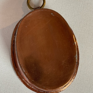 May include: A copper-colored, oval-shaped pan with a brass ring attached at the top. The pan has a slightly raised edge and a smooth interior. The metal has a warm, reddish-brown hue, with some signs of wear.