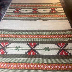 Può includere: Un tappeto tessuto con sfondo bianco e un motivo ripetuto di strisce rosse, verdi e marroni. Il tappeto ha un design geometrico con un motivo floreale.
