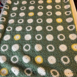 May include: Handmade crochet blanket featuring a daisy pattern. The blanket has a green background with white and yellow daisy motifs. The border is yellow. This cozy blanket is perfect for a bed or sofa.