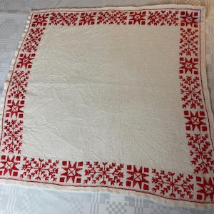May include: A square off-white textile cloth featuring a decorative border of red embroidered snowflake patterns. The central area of the fabric is plain and unadorned, while the edges are finished with a repeating geometric red stitch design.