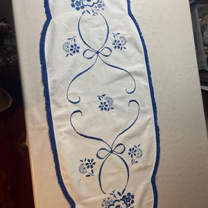 Op de afbeelding: Een langwerpige witte tafelloper met een blauw geborduurd bloemmotief met centrale strikken en gebogen lijnen. De loper heeft een geschulpte rand afgewerkt met een blauwe gehaakte bies, wat zorgt voor een decoratieve rand rondom.