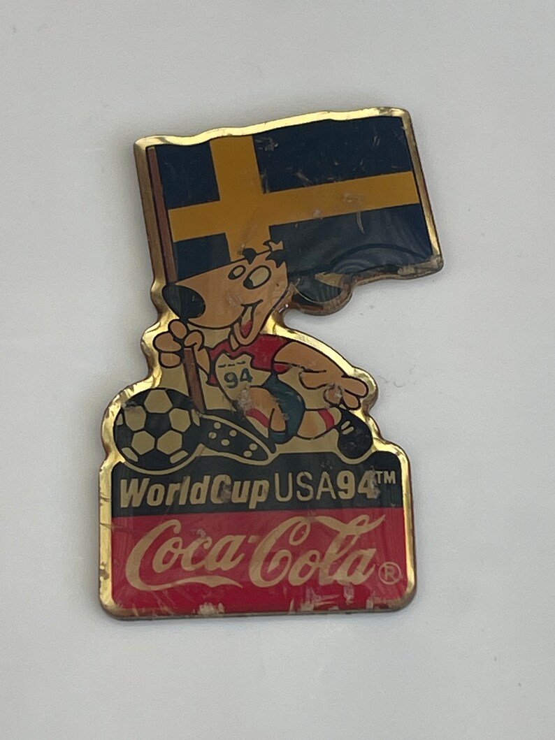 May include: Vintage pin featuring a cartoon dog holding a Swedish flag and a soccer ball. The pin has the text "World Cup USA94" and the Coca-Cola logo. The pin is gold-toned with red, black, and blue accents.