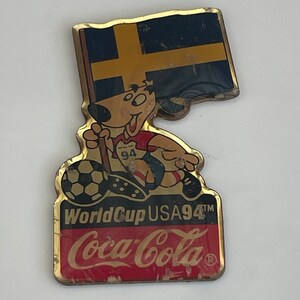 May include: Vintage pin featuring a cartoon dog holding a Swedish flag and a soccer ball. The pin has the text "World Cup USA94" and the Coca-Cola logo. The pin is gold-toned with red, black, and blue accents.