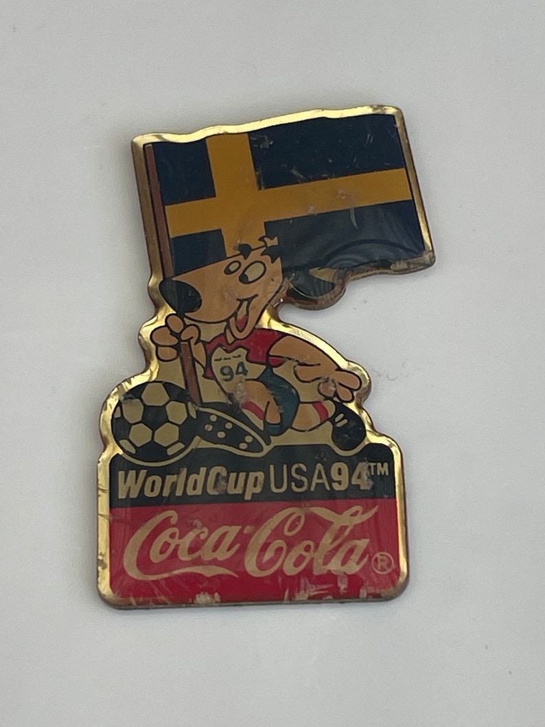 May include: Vintage pin featuring a cartoon dog holding a Swedish flag and a soccer ball. The pin has the text "World Cup USA94" and the Coca-Cola logo. The pin is gold-toned with red, black, and blue accents.