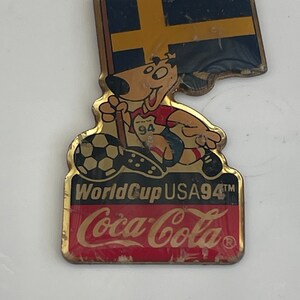 May include: Vintage Coca-Cola World Cup USA 94 pin. The pin features a cartoon dog wearing a soccer uniform and holding a soccer ball. The pin also includes the Swedish flag and the Coca-Cola logo.