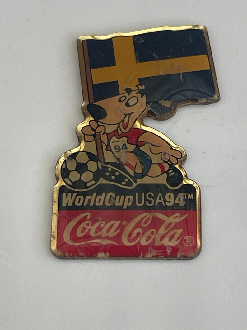 May include: Vintage Coca-Cola World Cup USA 94 pin. The pin features a cartoon dog wearing a soccer uniform and holding a soccer ball. The pin also includes the Swedish flag and the Coca-Cola logo.