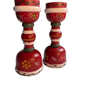 May include: Two red wooden candle holders with white, green, and yellow painted details. The candle holders have a floral design and are perfect for adding a touch of rustic charm to your home decor.