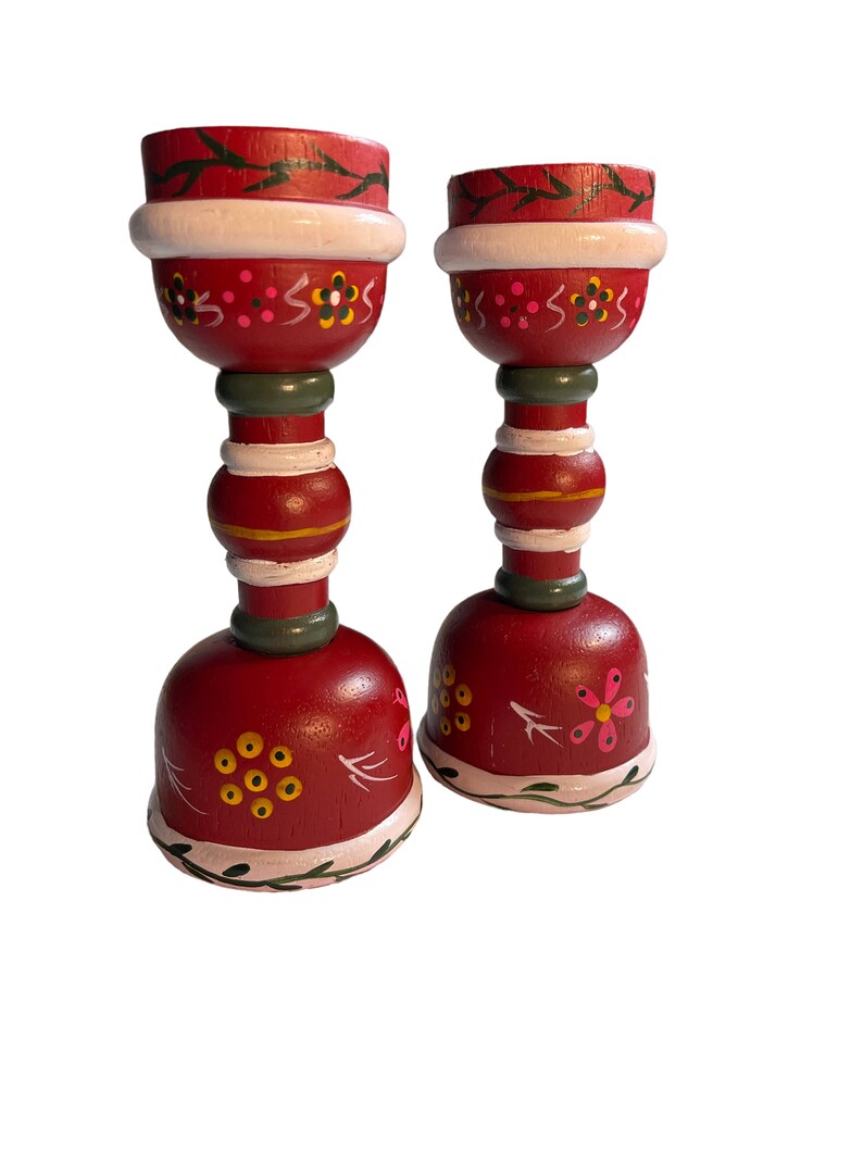 May include: Two red wooden candle holders with white, green, and yellow painted details. The candle holders have a floral design and are perfect for adding a touch of rustic charm to your home decor.