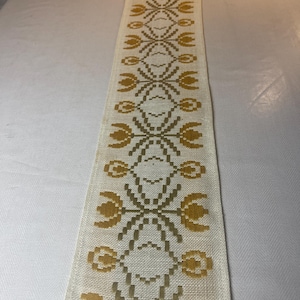 May include: A cream-coloured table runner with a repeating pattern of yellow-gold stylized flowers and geometric shapes. The runner is made of woven fabric and is displayed on a white surface.