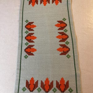 Op de afbeelding: Een rechthoekige, lichtblauwe geborduurde tafelloper met een groene rand. Het ontwerp heeft herhaalde oranje en bruine bloemmotieven met groene accenten. De loper is waarschijnlijk gemaakt van linnen of een vergelijkbare stof.