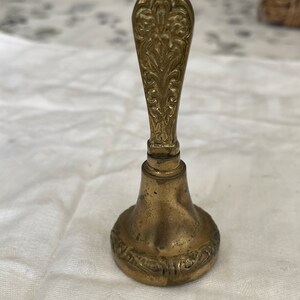 May include: A small, ornate brass bell with a detailed, floral-patterned handle. The bell has a rounded base and a slightly flared opening.