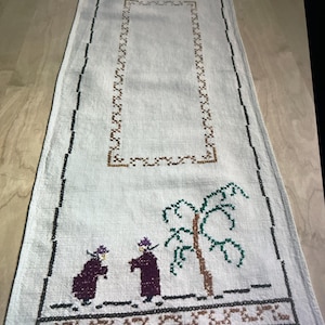 May include: White linen table runner with embroidered figures and a tree in brown, purple, green, and black thread. The runner has a brown and black border.