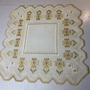 Peut inclure: Nappe brodée de couleur crème avec un centre carré et un bord festonné. La nappe présente des motifs floraux et géométriques en fil d'or et vert, créant un design délicat et orné.