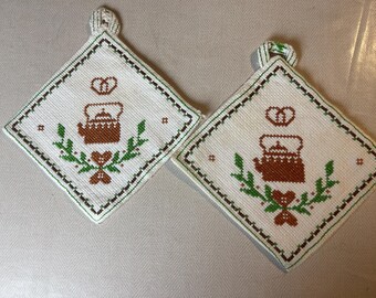 Könnte beinhalten: Zwei weiße Baumwoll-Topflappen mit brauner und grüner Stickerei einer Teekanne mit einem Herzen und Blättern. Die Topflappen haben oben eine Schlaufe zum Aufhängen.