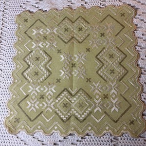 May include: A green embroidered table runner with a geometric pattern in white and brown thread. The runner has a scalloped edge.
