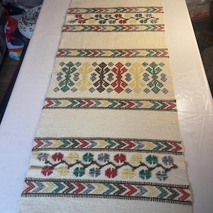 May include: A white woven table runner with a geometric pattern in shades of red, green, brown, and yellow. The runner has a fringe on both ends.