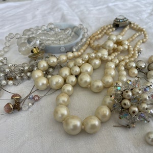 Pode incluir: Uma variedade de joias vintage, incluindo colares de pérolas, um broche de cristal e uma pulseira de contas transparentes. Os colares variam em tamanho e estilo, com pérolas pequenas e grandes. O broche apresenta cristais transparentes.