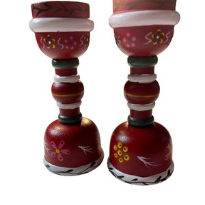 May include: A pair of red wooden candle holders with white, green, and yellow painted details. The candle holders have a bell-shaped base and a cylindrical top.
