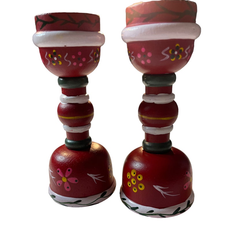 May include: A pair of red wooden candle holders with white, green, and yellow painted details. The candle holders have a bell-shaped base and a cylindrical top.