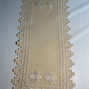 May include: A beige table runner with a white embroidered border and floral design. The runner has a delicate, lacy appearance.