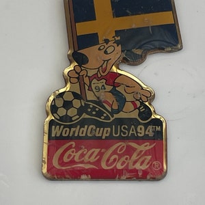 May include: Vintage Coca-Cola World Cup USA 94 pin. The pin features a cartoon dog wearing a soccer uniform and holding a soccer ball. The pin also includes the Swedish flag and the Coca-Cola logo.
