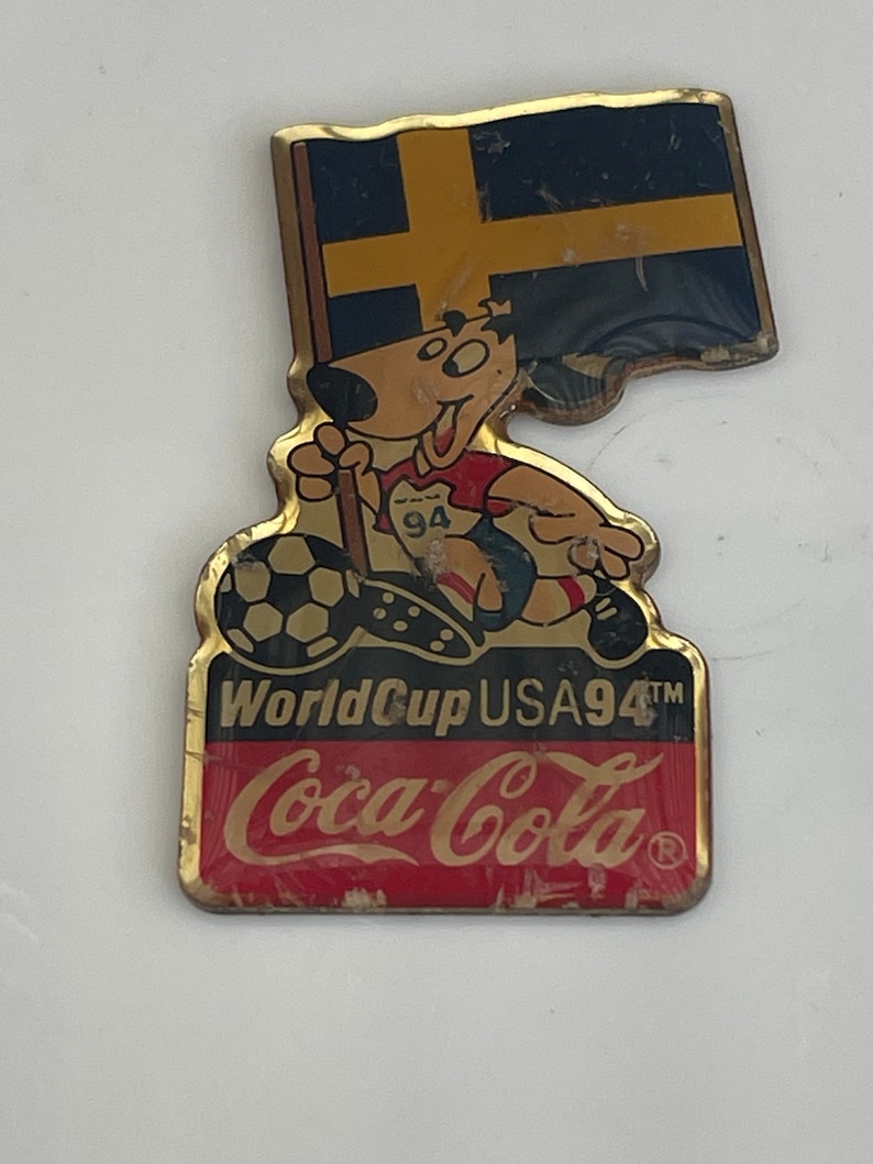 May include: Vintage Coca-Cola World Cup USA 94 pin. The pin features a cartoon dog wearing a soccer uniform and holding a soccer ball. The pin also includes the Swedish flag and the Coca-Cola logo.