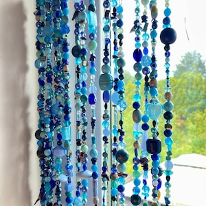 Puede incluir: Cortina de ventana hecha de hilos de cuentas azules en varios tonos. Las cuentas son de diferentes formas y tamaños, creando un efecto decorativo y texturizado. La cortina cuelga frente a una ventana, con vistas a vegetación.