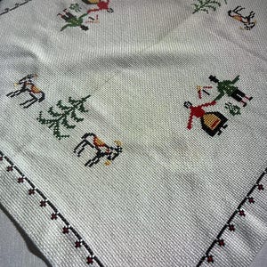 May include: White cotton table runner with a red, black, and green embroidered design of people, trees, and animals. The design is repeated on all four sides of the runner.