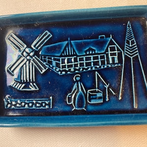 Pode incluir: Uma bandeja retangular de cerâmica com um esmalte azul profundo apresentando um design em relevo. A cena retrata um moinho de vento, uma casa, uma figura e uma estrutura alta. O acabamento brilhante destaca os detalhes em relevo no fundo.
