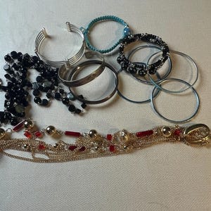 Op de afbeelding: Een verzameling armbanden en kettingen met verschillende materialen, waaronder zwarte kralen, zilver en goud. De sieraden zijn op een wit oppervlak gerangschikt.