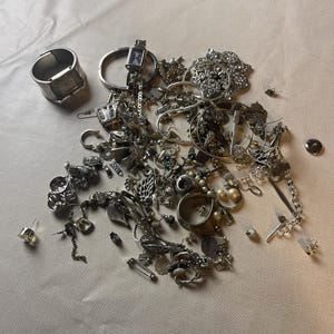 Può includere: Una collezione di gioielli color argento, tra cui orologi, collane, orecchini e anelli, è sparsa su una superficie di colore chiaro. L'assortimento presenta vari stili, dalle catene delicate ai pezzi importanti, mostrando una gamma di trame e design.
