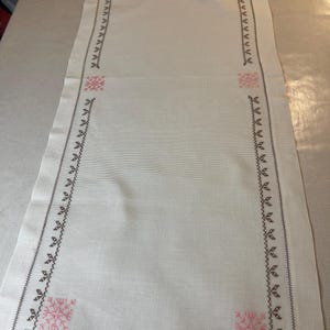 Op de afbeelding: Een rechthoekige witte tafelloper met een gedetailleerde geborduurde rand. De rand is donkerbruin met kleine bladachtige ontwerpen en roze bloemaccenten in elke hoek. De loper is gemaakt van een textuurstof.