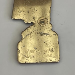 May include: A vintage brass-colored metal pin in the shape of a state, with a clasp on the back. The pin has a worn appearance with scratches and the text "&copy;1991 WC 94 MADE IN TAIWAN".