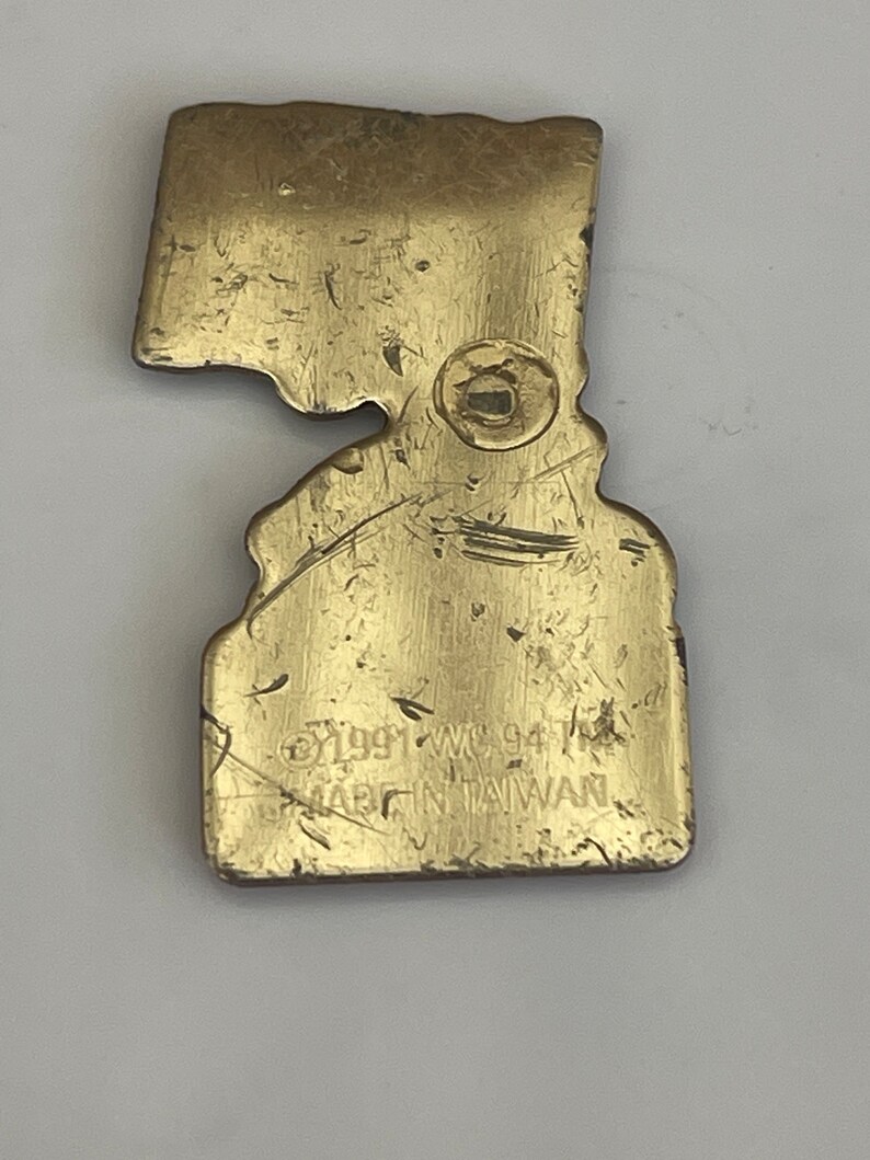 May include: A vintage brass-colored metal pin in the shape of a state, with a clasp on the back. The pin has a worn appearance with scratches and the text "&copy;1991 WC 94 MADE IN TAIWAN".