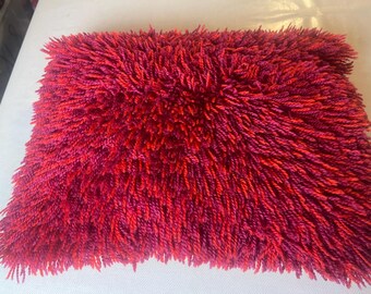 May include: A rectangular decorative pillow with a shaggy, textured surface. The pillow's color transitions from a deep red to a vibrant magenta, creating a visually appealing gradient. The plush texture suggests a soft and comfortable feel, suitable for home decor.
