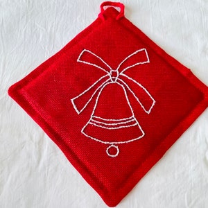 Può includere: Un sottopentola rosso a forma di diamante con un disegno di campana ricamata bianca. Il sottopentola ha un anello per appenderlo ed è realizzato in tessuto strutturato. La campana ha un fiocco in cima.
