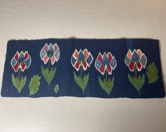 May include: A dark blue woven rug with a repeating pattern of colorful flowers. The flowers are stylized and have a geometric design. The rug is approximately 12 inches wide and 36 inches long.