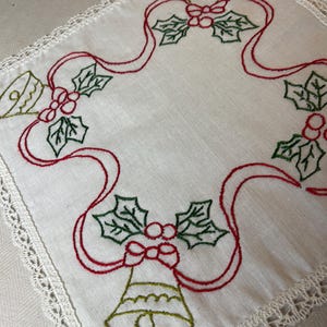May include: A square white fabric napkin featuring an embroidered border design of green bells, red ribbons, and green holly leaves with red berries. The edges of the napkin are finished with a delicate white lace trim along all four sides.