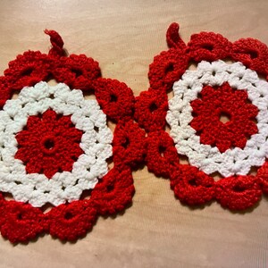 Könnte beinhalten: Zwei handgefertigte Topflappen aus Häkelarbeit. Jeder Topflappen hat ein rot-weißes Blumendesign. Die Mitte ist rot, umgeben von einem weißen Ring und einem roten, gewellten Rand. Eine Schlaufe zum Aufhängen ist angebracht.