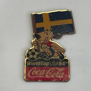 May include: A vintage Coca-Cola pin featuring a cartoon dog holding a soccer ball and a Swedish flag. The pin is gold-toned with the text "World Cup USA94" and the Coca-Cola logo in red.
