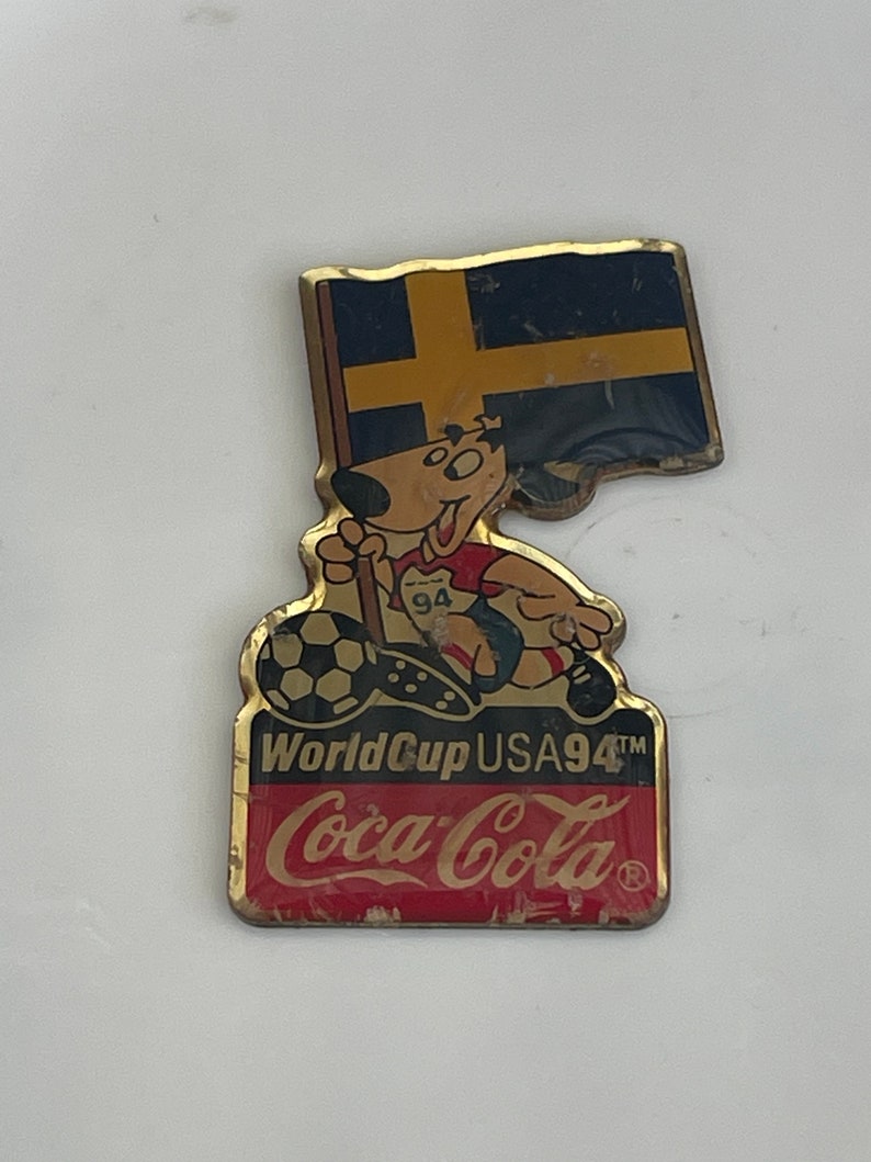 May include: A vintage Coca-Cola pin featuring a cartoon dog holding a soccer ball and a Swedish flag. The pin is gold-toned with the text "World Cup USA94" and the Coca-Cola logo in red.