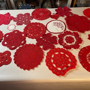 Könnte beinhalten: Eine Sammlung verschiedener roter Häkeldeckchen in unterschiedlichen runden, floralen und geometrischen Formen. Ein weißes rundes Deckchen weist ein rotes gesticktes Blumenmuster auf. Die Artikel sind in einer Gruppe angeordnet und zeigen filigrane Handarbeit.