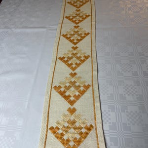 May include: A long, rectangular table runner made of off-white fabric featuring a series of repeating diamond-shaped patterns embroidered in shades of golden yellow and orange. The runner has a stitched border in a matching golden yellow thread.