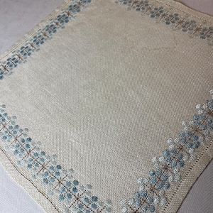 May include: A square beige linen napkin featuring a delicate embroidered border of small blue and light brown floral patterns. The embroidery follows the perimeter of the fabric, creating a decorative frame around the plain center of the textile.