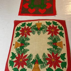 May include: Two square Christmas placemats. One features a brown center with red hearts and a green square design, framed by a red border. The other has a beige center with a wreath of red poinsettias, green leaves, and gold bells, with a red border.