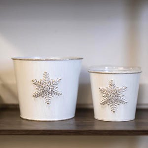 Pode incluir: Dois baldes de metal branco com desenhos de flocos de neve prateados, um grande e outro pequeno, estão numa prateleira de madeira escura. Uma pinha está à esquerda. Os baldes têm um acabamento envelhecido e uma borda metálica.