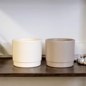 May include: Two ceramic plant pots, one cream and the other tan, are displayed on a dark wooden shelf. The pots have a simple, modern cylindrical design with a small base. A decorative beaded strand is also on the shelf.