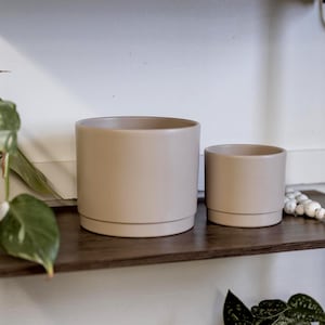 May include: Two beige ceramic plant pots of different sizes sit on a dark brown wooden shelf. The pots are cylindrical with a matching saucer base. Other plants and pots are visible on the shelves above.