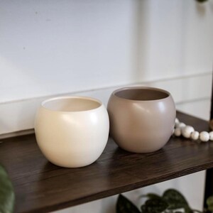 May include: Two small, round ceramic planters in cream and taupe sit on a dark wood shelf. The planters have a smooth, matte finish and a wide opening. The shelf is part of a black metal shelving unit, with greenery in the foreground.