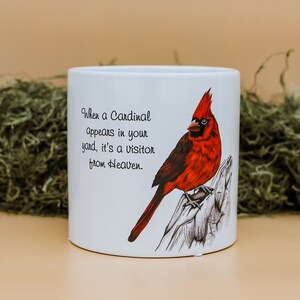 May include: White ceramic mug with a detailed illustration of a red cardinal on a branch. The mug features the text: "When a Cardinal appears in your yard, it's a visitor from Heaven." The background is a beige colour.
